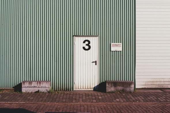 This is a photo of a white door with the number 3 on it, set into a building with a corrugated green facade. The door is flanked by a mailbox with multiple slots and labels on the right side, indicating it's likely a shared or multi-unit building. There's a grey concrete block on the left side of the door, perhaps a step or a place to rest deliveries. The ground in front of the door is paved with bricks.