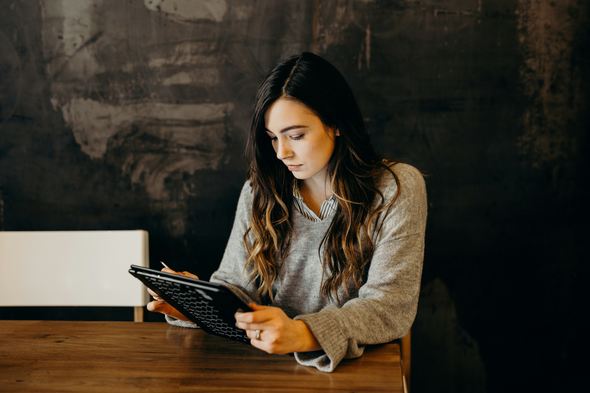 A woman with long dark hair, wearing a gray sweater, is sitting at a wooden table, focused on a tablet with a keyboard. The background is dark with a textured surface, creating a cozy and quiet atmosphere.